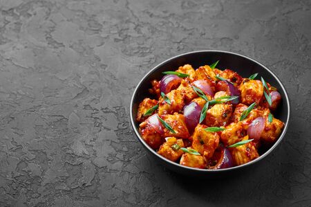 Schezwan Paneer in black bowl at dark slate background. Schezwan Paneer is indo-chinese cuisine dish with deep fried Paneer cheese, onion and Schezwan Sauce. Copy Spaceの写真素材