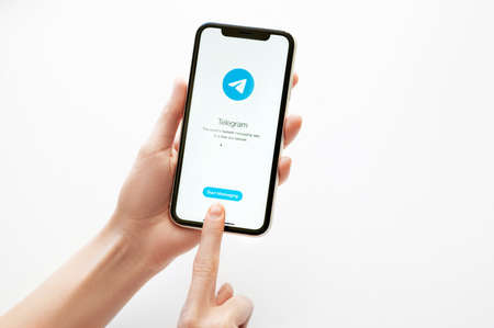Moscow, Russia, 28 April, 2020. Telegram application logo on screen of Iphone X on white backdrop. Telegram messenger app icon on smartphone display. Female hands keeps a phoneのeditorial素材
