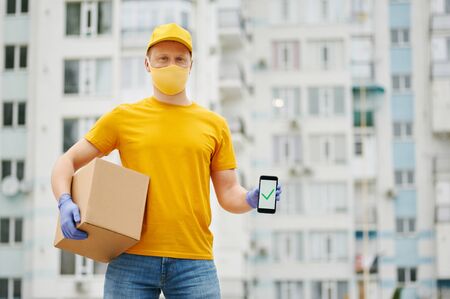 Delivery Man employee in yellow uniform cap, t-shirt, face mask and gloves holds a cardboard box package shows complete sign on the phone. Safety delivery quarantine service in covid-19 virus pandemicの写真素材
