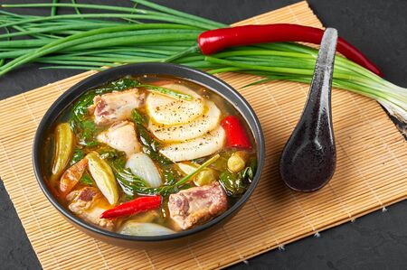 Sinigang na Baboy or Filipino Pork Meat Soup in black bowl on dark slate backdrop. Sinigang is a Filipino cuisine dish with meat, bamia, daikon, spinach, fish sauce. Filipino Food. Asian Mealの写真素材