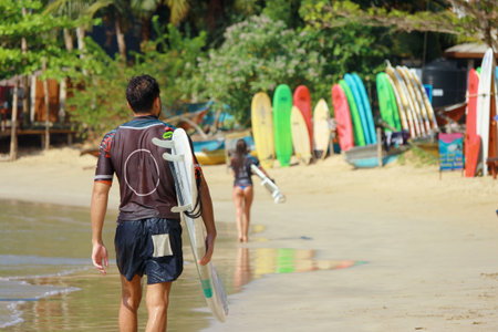 Back view of a surfer man walking by the tropical beach carrying a surfboard.