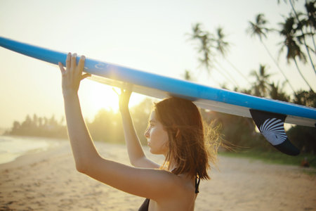 A young surfer woman stands on the tropical beach with a blue surfboard before surfing at sunset