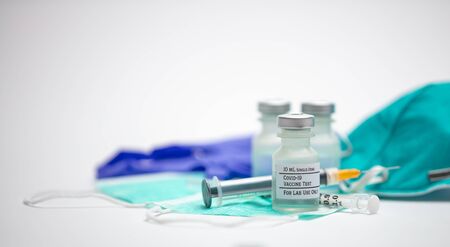 Covid-19 vaccine test vials on top of medical face masks.  Syringes, rubber medical gloves and respirator mask in background.の写真素材