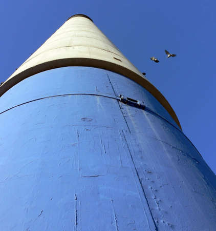 Shot of a blue, white and black chimney with pigeons flying away from it, from the bottom up, against a blue sky.の写真素材