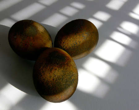 Three eggs which have been treated with a rust paint finish, against a white tabletop with patterns of shadow and light across all.の写真素材