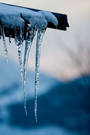 Icicles hang off a roof; one in sharp focus, at dusk, with vague landscape background.の写真素材