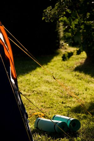 An orange tent, with green or blue rolled-up bedrolls beside it, in a meadow in the evening light.の写真素材