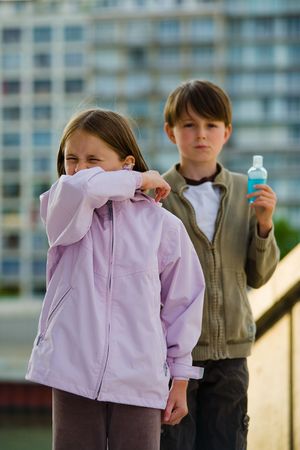 Two children stand in an urban setting, one sneezing into their elbow, the other holding a bottle of soapless hand cleanser.の写真素材