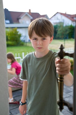 A boy in a green t-shirt stands in an open door with an angry, troubled expression on his face, while his sister appears indistinctly in the background.  The brother and sister have been arguing, and he is angry.の写真素材