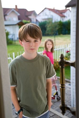 A boy in a green t-shirt stands in an open door with an angry, troubled expression on his face, while his sister appears indistinctly in the background.  The brother and sister have been arguing, and he is angry.の写真素材