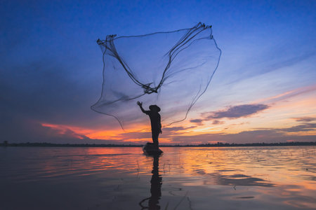 Asian fisherman on wooden boat casting a net for catching freshwater fish in natureの写真素材