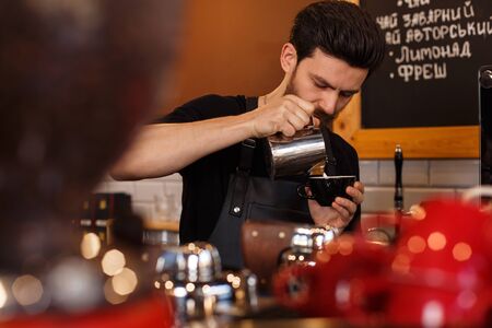 Young smiling male barista making cappuccino. Pouring milk for prepare cup of coffee. Latte art. Morning cup of coffee in cafÃ©. Brewing coffee. Coffee shop concept.の写真素材