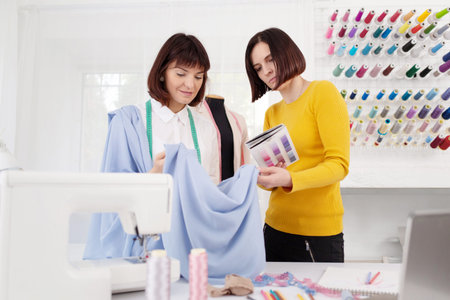Two young female dressmakers working in sewing studio. Seamstress select color of threads suitable...