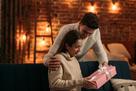 Handsome man presenting a gift to his beautiful girlfriend and smiling. Beautiful young couple at home enjoying spending time together.Valentines Day, relationships and people concept.の写真素材