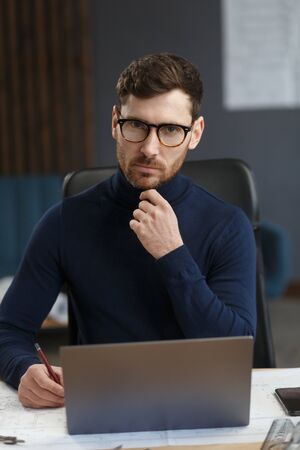 Young businessman working in office with laptop. Business portrait of handsome bearded man wearing eyeglasses sitting at workplace. Confident businessman became successful. Business concept.の写真素材