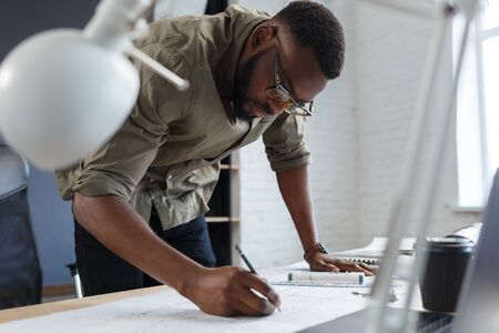 Afro-American architect working in office with blueprints.Engineer inspect architectural plan, sketching a construction project. Portrait of black handsome man sitting at workplace. Business concept.の写真素材
