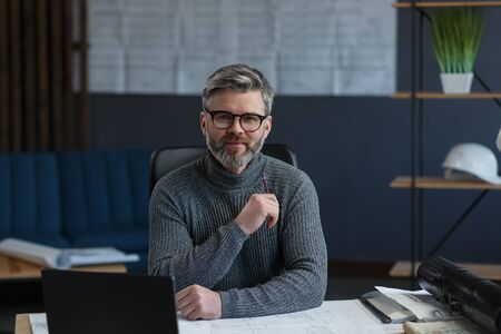 Business portrait of handsome bearded man wearing eyeglasses sitting at workplace. Confident businessman became successful. Interior designer working in office with laptop. Business concept.の写真素材