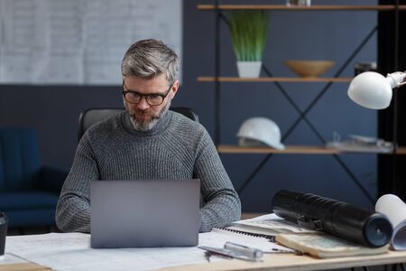 Designer working in office with laptop. Architect thinks over architectural plan, searching new ideas for construction project. Portrait of handsome bearded man sitting at workplace. Business concept.の写真素材