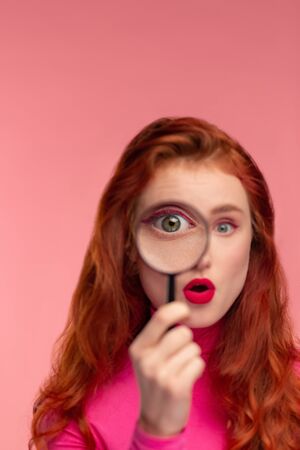 Young detective. Close-up portrait of surprised redhead girl looking at camera through magnifying glass, over pink background. Funny model with long hair starting investigation. Womans intuition.の写真素材