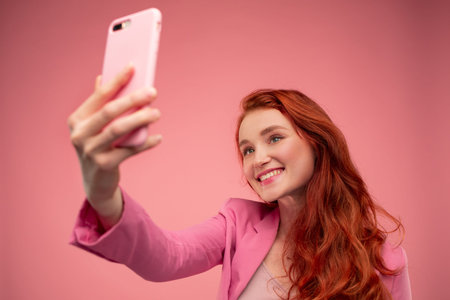 Beautiful young redhead woman making selfie. Funny smiling girl wearing pink jacket holding pink...