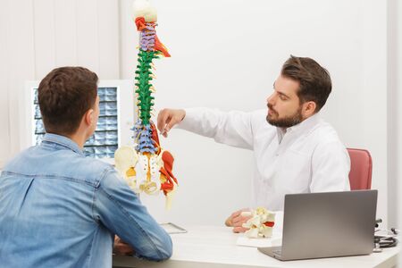 Doctor showing flexible spine model to his patient and explains the cause of his pain in medical office. Patient visiting physiotherapist in clinic. Surgery, healthcare and medicine concept.の写真素材