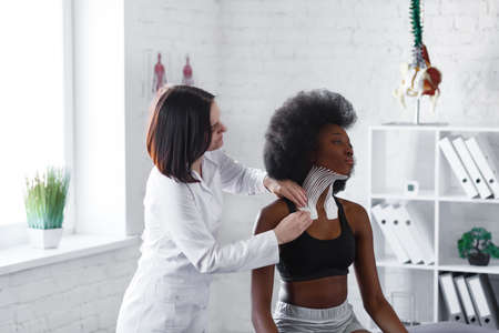 Kinesiology taping. Physiotherapist applying kinesiology tape to patient neck. Therapist treating young female African American athlete. Post traumatic rehabilitation, sport physical therapy.の写真素材
