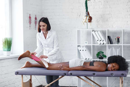 Kinesiology taping. Physiotherapist applying kinesiology tape to patient calf muscle.Therapist treating young female African American athlete. Post traumatic rehabilitation, sport physical therapy.の写真素材