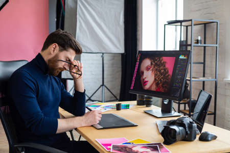 Portrait of tired graphic designer working overtime in office. Stressed worker have eye strain symptoms. Retoucher workplace in photo studio. Burnout and overwork concept. Creative agency.の写真素材