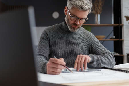 Architect drawing blueprints in office. Engineer sketching a construction project. Architectural plan. Close-up portrait of handsome bearded man concentrated on work. Business construction concept.の写真素材