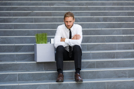 An unhappy young manager in a white shirt is fired from his job. A sad worker sits on the steps of a...