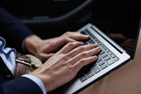 Close-up photo of male hands with laptop. Businessman in suit sitting in car, texting message, searching web, browsing information using laptop keyboard. Online education, remote working.の写真素材