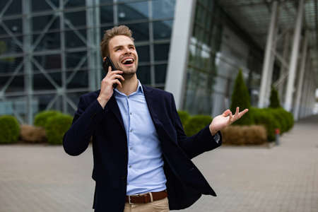 Portrait of handsome smiling man in casual wear holding smartphone. Successful manager makes a call. Young businessman talking on the phone, standing on city street near business center.の写真素材