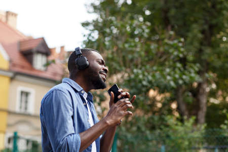 Walking in the street, relaxation, leisure. Young Afro-American man in headphones listening music on smart phone using music app. Portrait of smiling guy in earphones and mobile phone outdoors.の写真素材