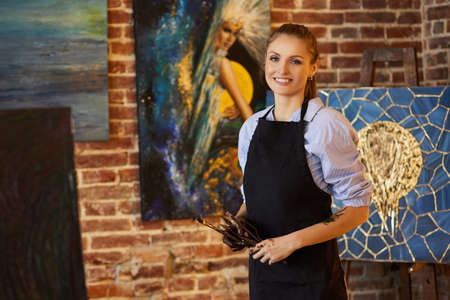 Portrait of young smiling female artist wearing apron in art studio. Painter holding art brushes, hands are stained with paint. Creative process, relaxation, leisure, hobby, stress management.の写真素材