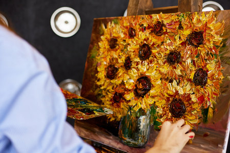 Young beautiful female artist painting still life with sunflower on canvas using oil paintings and palette knife. Painter creating artwork in art studio. Relaxation, leisure, hobby, stress managementの写真素材