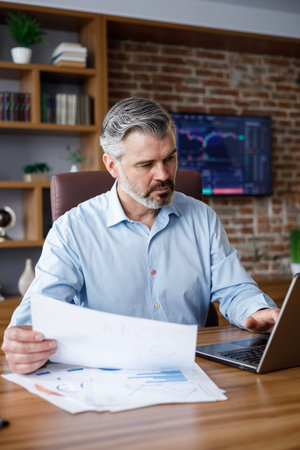 Portrait of adult gray-haired bearded businessman working with laptop at home office. Texting...