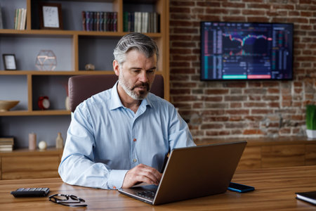 Portrait of adult gray-haired bearded businessman working with laptop at home office. Texting...
