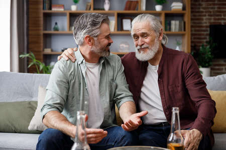 Fathers day. Adult son with senior father drinking beer sitting on couch at home. Two generations family enjoy pleasant conversation, talking, sharing news, spending time togetherの写真素材