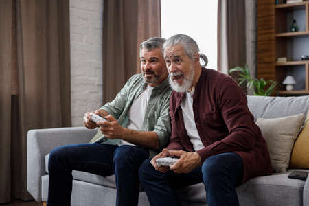 Adult son and senior father having fun, enjoy spending time together. Two generations family playing video games using gamepads at home. Fathers Day. Active modern elderly peopleの写真素材