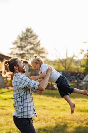 Happy little kid having fun with dad in domestic garden on warm sunny day. Father throwing his child in the air. Spending time together on weekend at the countryside. Weekend with dad. A happy familyの写真素材