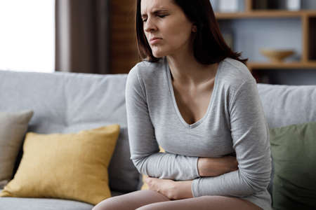 Sick young woman suffering from menstrual pain. Woman with hands squeezing belly having painful stomach ache or period cramps sitting on sofa, Abdominal pain, gastritis, and painful periods conceptの写真素材