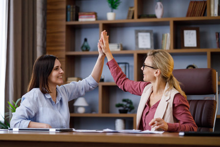 Two young smiling business women giving high-five in modern office. Happy female workers celebrating...