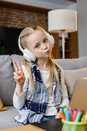 Portrait of cute primary school girl studying at home using laptop computer. Smiling child wearing white earphones working on computer. Distance learning. Children home educationの写真素材
