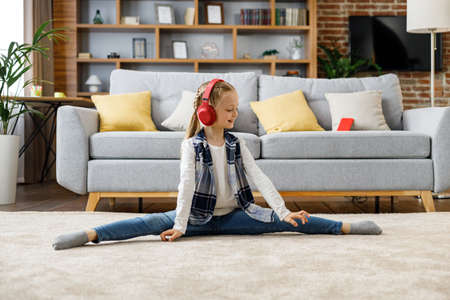 Smiling sporty little girl sitting on the twine raising on carpet at home. Home training of a little ballerina. Active healthy leisure for kidsの写真素材