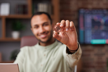 Portrait of smiling businessman holding bitcoin in office interior with stock market graphs on...