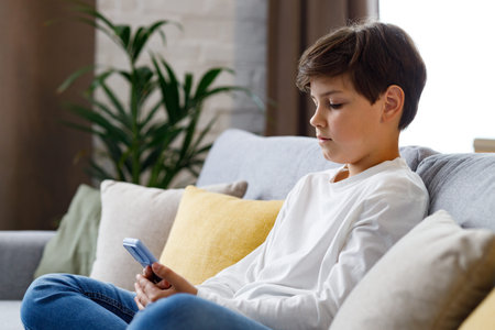 Portrait of young cute boy holding smartphone sitting at sofa. Teenager at home choosing favorite...