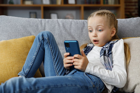 Surprised little cute girl holding smartphone and resting at sofa at home. Spending time on social networks, texting messages,browsing internet,watching cartoons, playing games on mobile phoneの写真素材