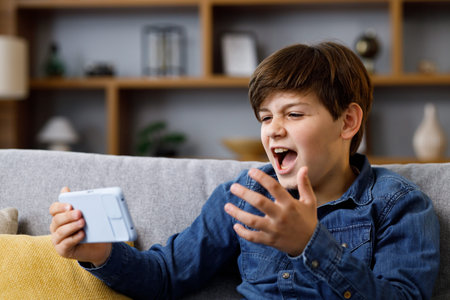 Young boy reacts emotionally to what is happening on the smartphone screen. Teenager spending time at home with digital gadget. Adolescence and pubertyの写真素材