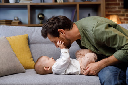 Dad plays with his little baby, on the couch in a cozy apartment. Fatherly love. Father hugs and kisses his little daughter.の写真素材