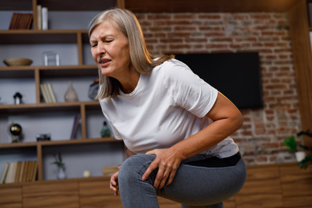 Gray-haired mature woman doing gymnastics and yoga at home and gets a knee injury. Pain in the knee, cramp. Injuries in fitness, stretch marks. Well-being of the elderly.の写真素材